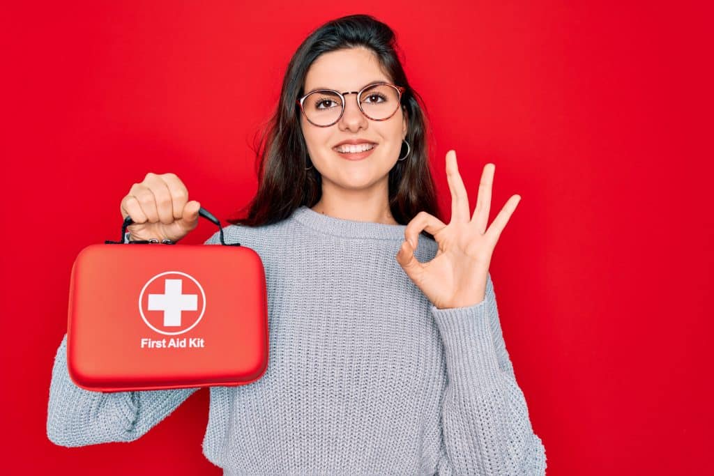 Junge Frau mit Brille zeigt einen roten Erste-Hilfe-Kasten und hebt ihre Hand zum OK-Zeichen.
