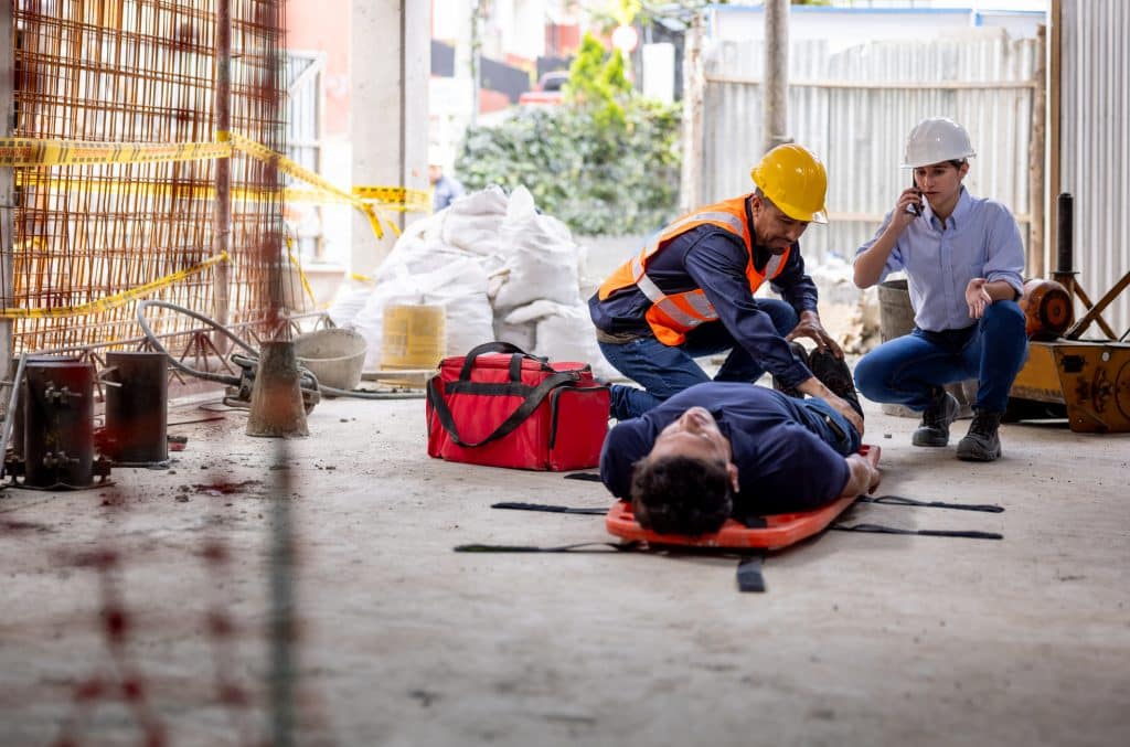 Zwei Mitarbeitende stabilisieren eine verletzte Person auf einer Trage in einer Baustelle. Daneben liegt eine rote Erste-Hilfe-Tasche.
