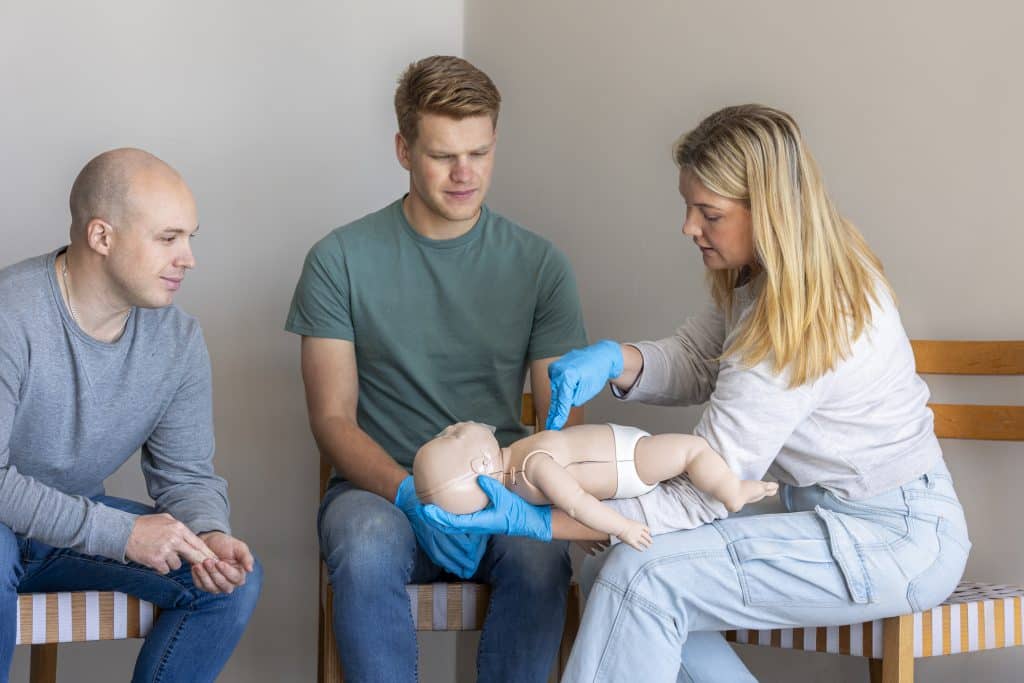 Ausbilderin demonstriert an einer Baby-CPR-Puppe die richtige Handposition zur Ersten Hilfe, während zwei Teilnehmende zusehen.